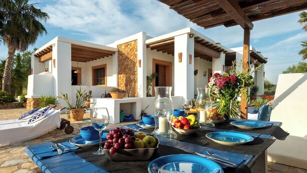 Un espacio de comedor al aire libre con un muro de piedra, una pérgola de madera y una mesa dispuesta con platos azules, vasos y un cuenco de fruta.