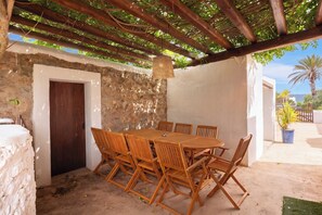 Zona de menjador a l'aire lliure coberta, amb paret de pedra, taula i cadires de fusta.