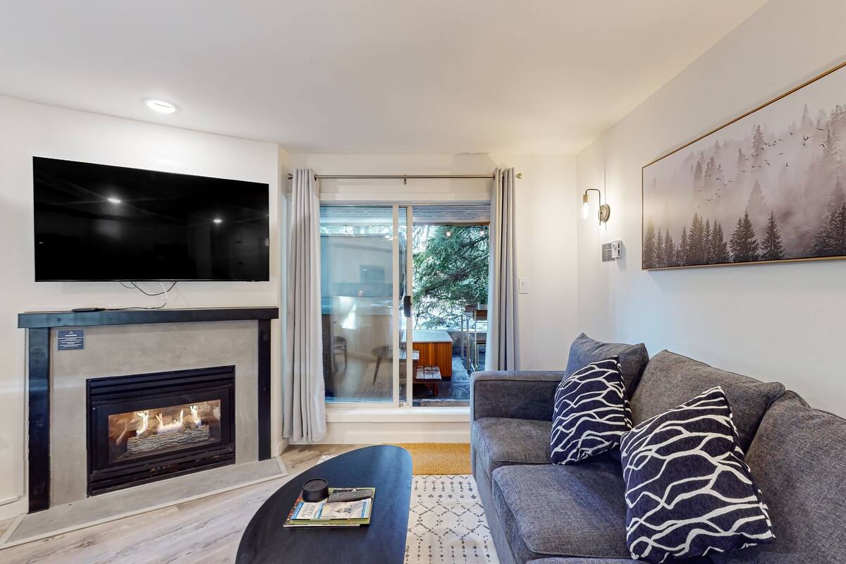 Ein modernes Wohnzimmer mit Kamin, Flachbildfernseher, grauem Sofa mit gemusterten Kissen und einem großen Fenster mit Blick ins Freie.