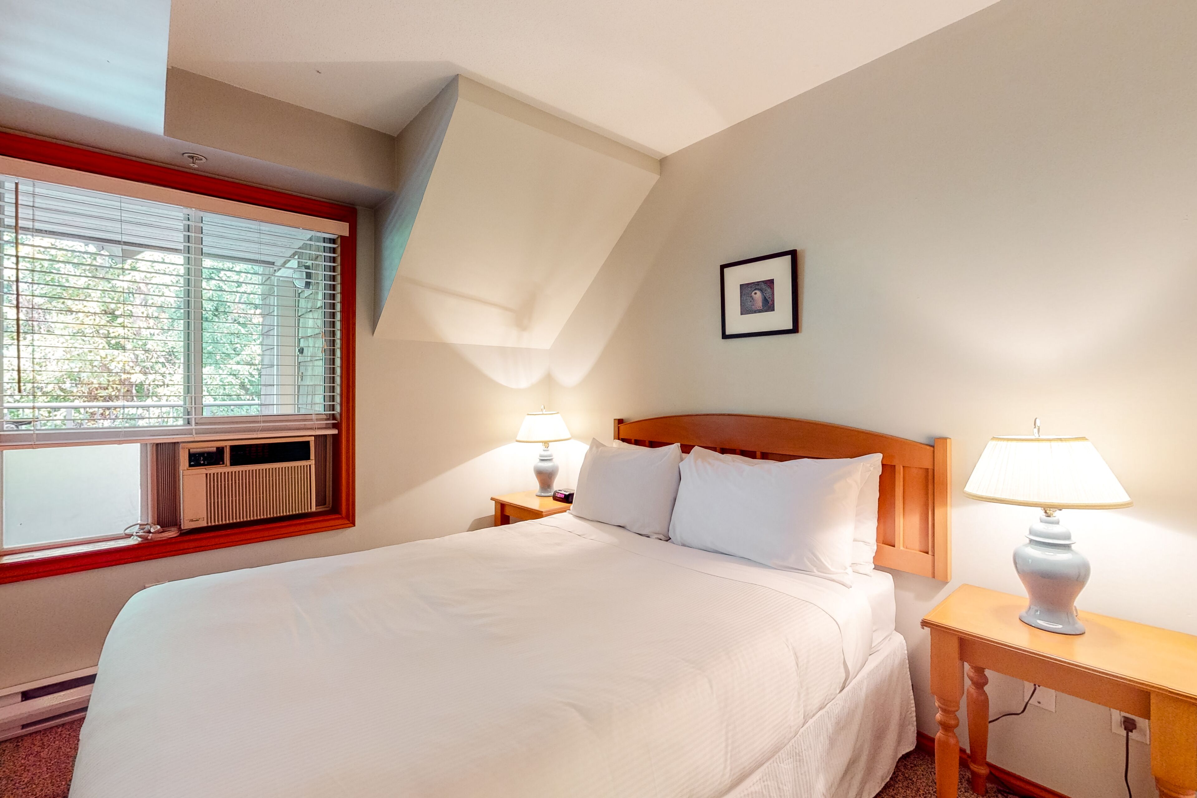Habitación de hotel con una cama grande, dos mesitas de noche con lámparas, una ventana con persianas, un radiador y un cuadro en la pared.