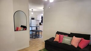 A living room with a black sofa, a mirror, and a kitchen in the background.