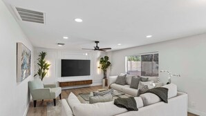 Una sala de estar moderna con sofá, sillón, televisor, ventilador de techo y ventanas amplias.