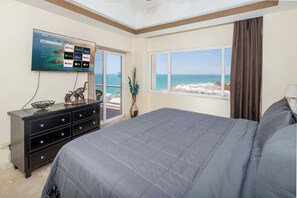 Ein Schlafzimmer mit Bett, Kommode, Fernseher der Streaming-Dienste anzeigt und einem Fenster mit Blick auf den Strand sowie einem Vorhang.