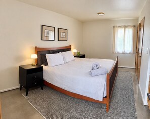 A neatly made bed with a white comforter and pillows, a bedside table with a lamp, a framed picture on the wall, and a window with curtains. - Ranch Retreat near Santa Fe • Indoor Pool, Hot Tub & Big Skies (Lamy)
