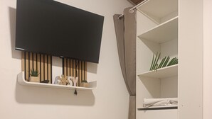 Ein Zimmer mit an der Wand montiertem Fernseher, einem Holzregal mit Dekoration, einem weißen Kleiderschrank und einer Ablage mit Blumen und Tasse auf dem Boden.