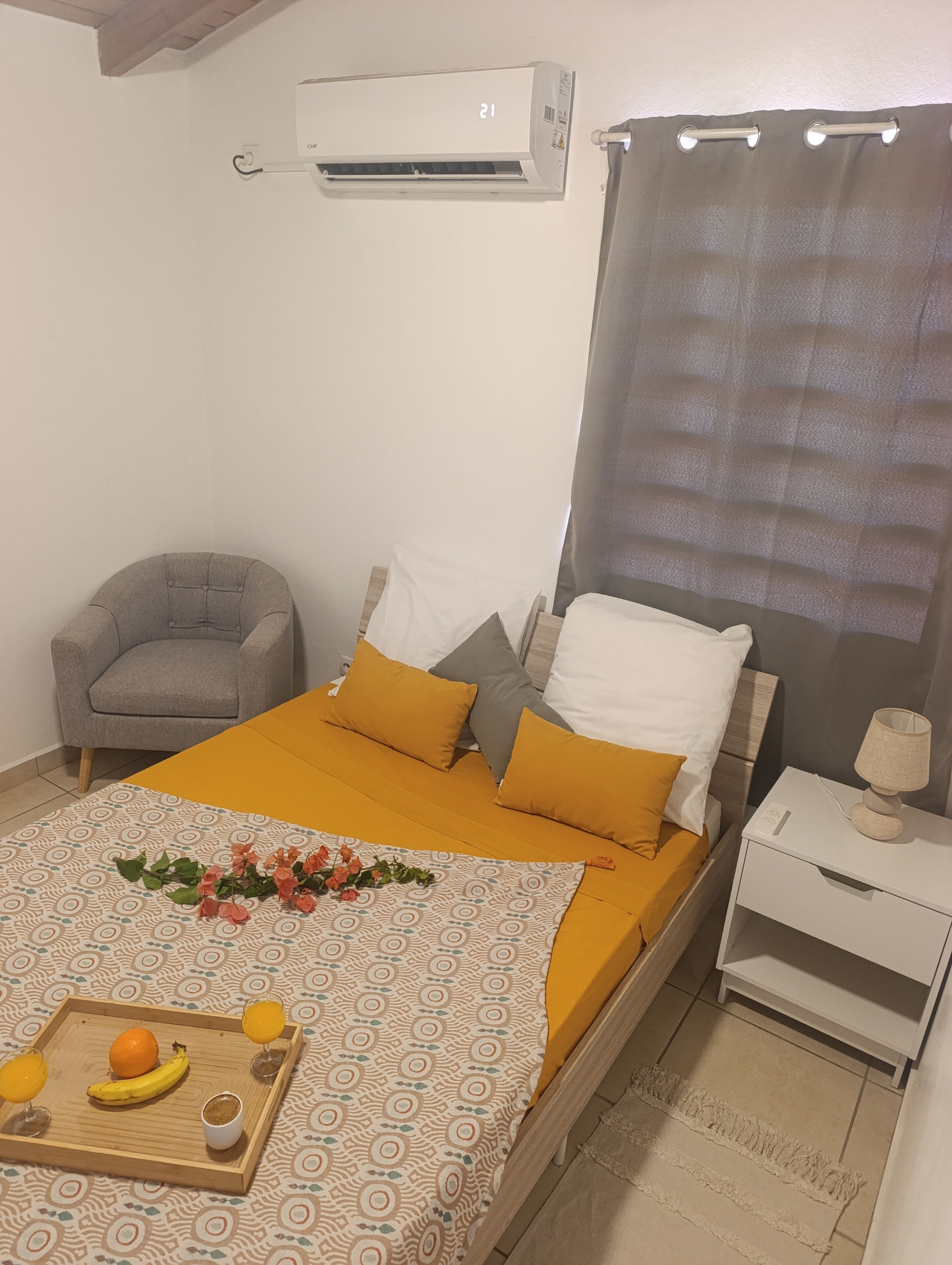 Un dormitorio ordenado con cama, una silla, una mesita de noche con lámpara y una bandeja con fruta y una bebida.