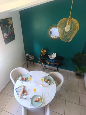 A dining area with a round table set for breakfast, a teal accent wall, a hanging light fixture, and a framed artwork on the wall. - Les hauts de Carrère, fleur-de-lys. (Petit-Bourg)