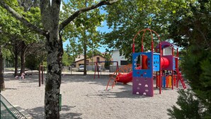 Un parque infantil con tobogán y estructuras para escalar, rodeado de árboles y un edificio al fondo.