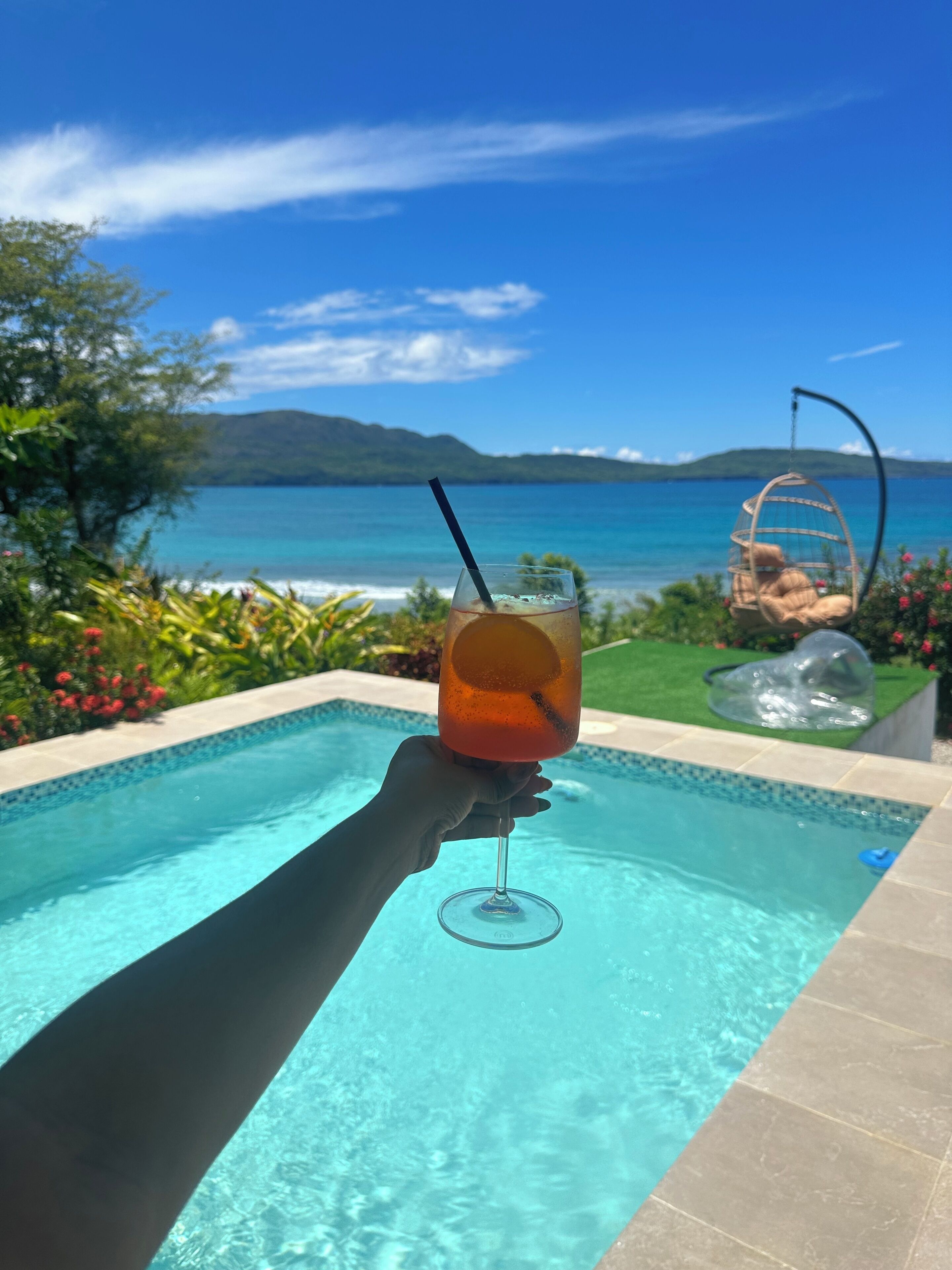 Una mano sostiene un vaso con una bebida junto a una piscina con vista al mar y las montañas.