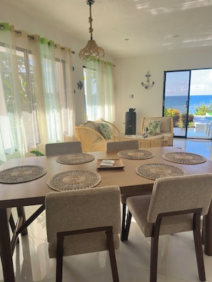 A dining area with a table set for six, surrounded by chairs, a sofa, and a view of the ocean through a sliding glass door. - Beachfront villa in Samaná with private pool (Las Galeras)
