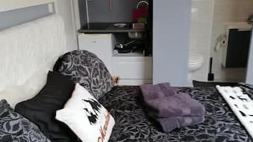 Een slaapkamer met een bed, kussens, een nachtkastje, een kleine kitchenette en een badkamer.