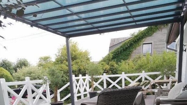 A covered outdoor deck with wicker furniture, a white fence, and a house in the background. - Le Domaine d'Eva (Libramont-Chevigny)