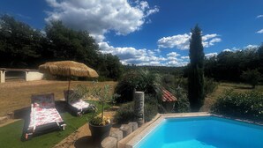 En swimmingpool omgiven av en trädgård med ett parasoll av halm, ett trädäck och krukväxter.