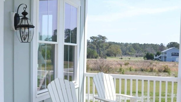 A covered porch with two Adirondack chairs, a white railing, and a view of a landscape. - Coastal Retreat Near Beaufort (Beaufort)