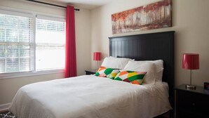 Un dormitorio con cama, ventana con persianas, cortina roja, un cuadro en la pared y dos lámparas de noche.