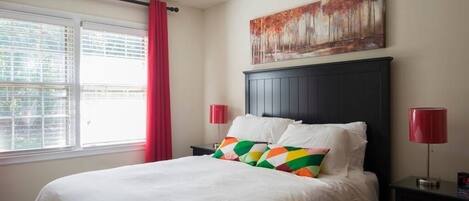 A bedroom with a bed, a window with blinds, a red curtain, a painting on the wall, and two bedside lamps.