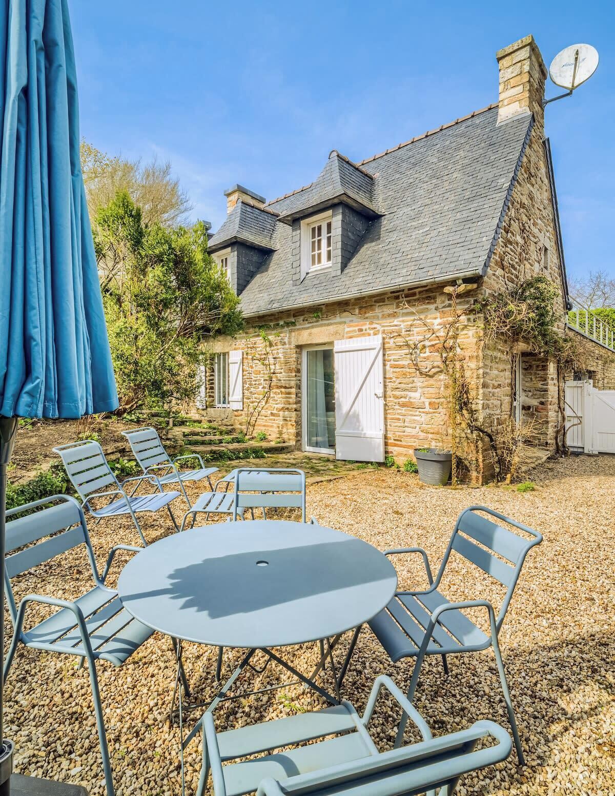 Une maison en pierre avec un toit en tuiles, une antenne parabolique et une terrasse en gravier avec des chaises et une table en métal.