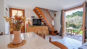 Ein Essbereich mit Tisch, Stühlen, einem Blumenstrauß, einer Holztreppe, einem Fernseher und Blick ins Freie.