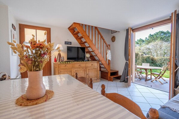 Un comedor con una mesa, sillas, un jarrón de flores, una escalera de madera, un televisor y vistas al exterior.