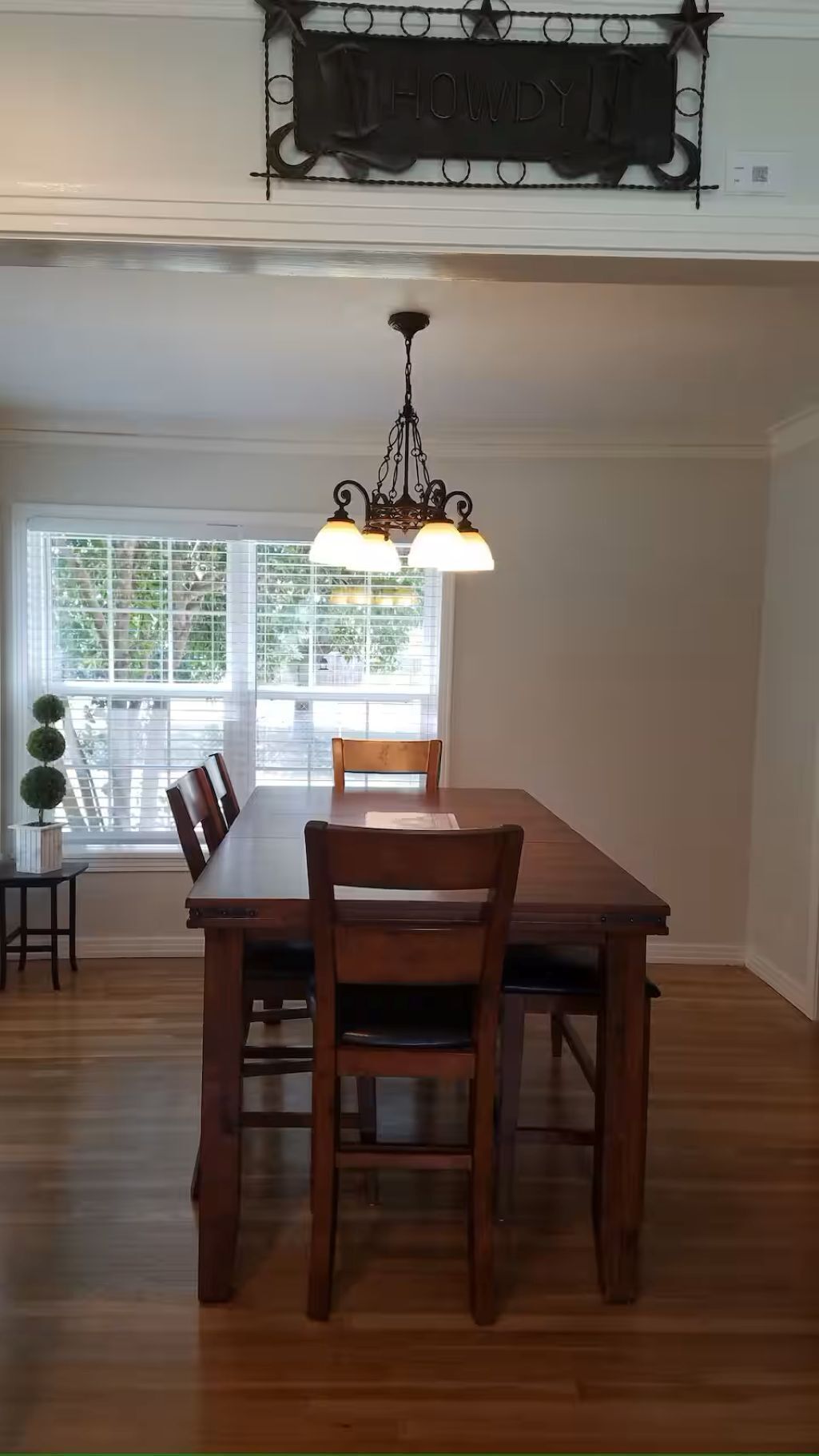 A dining area with a wooden table and chairs, a chandelier, and a window with blinds.