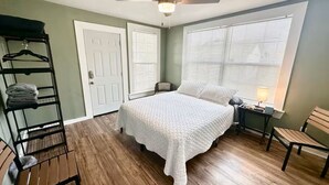 Quarto com cama, ventilador de teto, janela com persianas, mesa de cabeceira, luminária, banco, cadeira e cabideiro com roupas dobradas.
