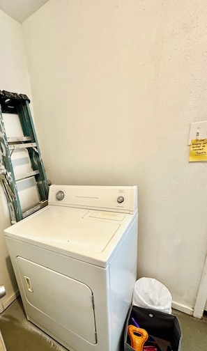Een wasserette met een witte wasmachine, een metalen plank en een prullenbak.
