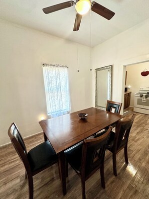 A dining area with a wooden table and chairs, a ceiling fan, a window with curtains, and a kitchen in the background. - Sleeps 4/ Fenced Yard/ Pet Friendly/ Near Downtown (San Angelo)