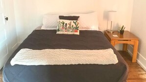 A bed with a dark mattress, a white blanket, and a decorative pillow. There is a wooden nightstand with a lamp and a small potted plant beside the bed. - Sleeps 4/ Fenced Yard/ Pet Friendly/ Near Downtown (San Angelo)