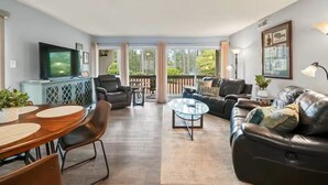 A spacious living room with a large sofa, a coffee table, and a TV. There is a balcony with a railing and a view of trees. - Birdie Bungalow - Sleeps 8 - Prime Location (Pinehurst)