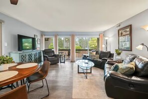 A spacious living room with a large sofa, a coffee table, and a TV. There is a balcony with a railing and a view of trees. - Birdie Bungalow - Sleeps 8 - Prime Location (Pinehurst)