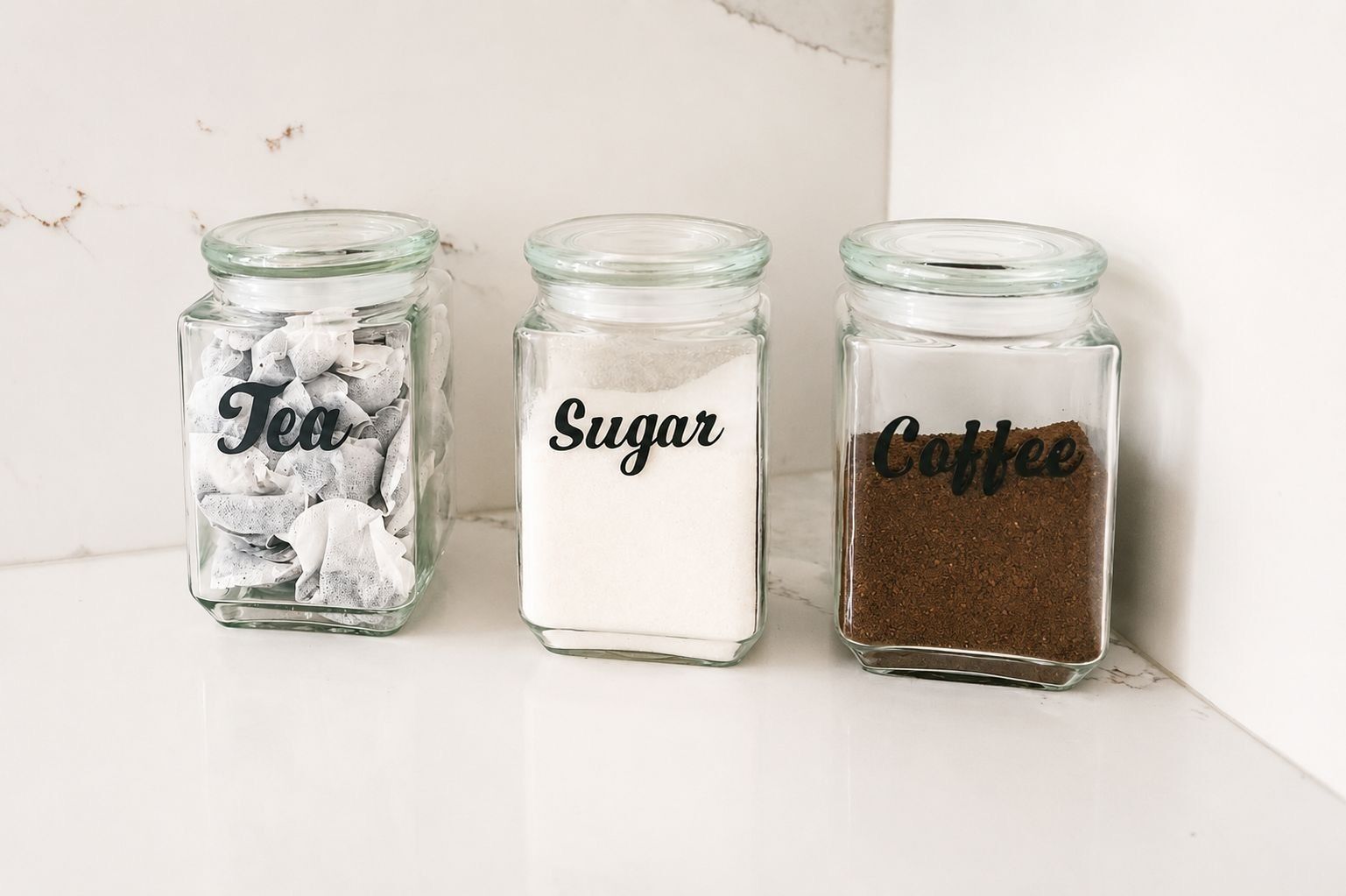 Tres frascos de vidrio etiquetados 'Té', 'Azúcar' y 'Café' sobre una superficie blanca.