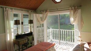A bedroom with a bed, a desk, a chair, a ceiling fan, and a view of a garden through a sliding door.