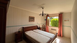 Quarto com cama, mesas de cabeceira, uma cadeira, janela com cortinas, um quadro na parede e um ventilador de teto.