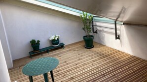 Un petit balcon doté d’un plancher en bois, de deux plantes en pot, d’une petite table et d’un banc.