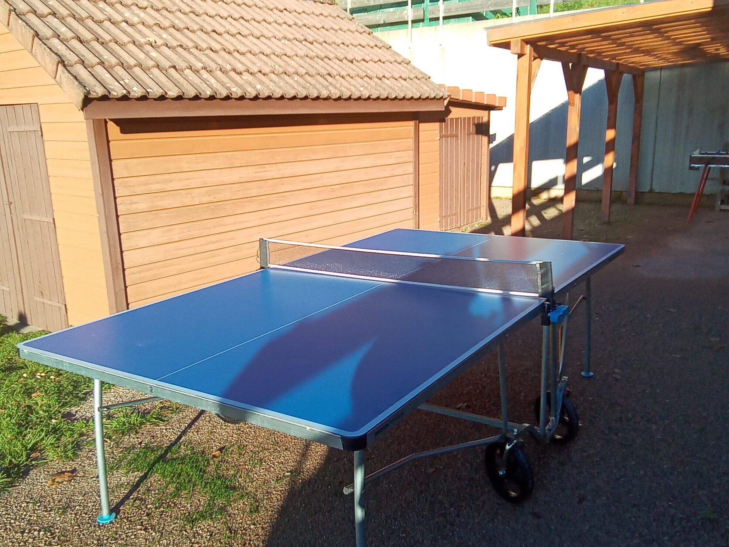 Una mesa de ping pong portátil al aire libre, con una estructura de madera al fondo.