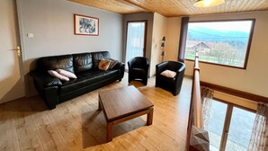 A living room with a wooden ceiling, a black leather sofa, a wooden coffee table, and a view of a rural landscape through a large window. - Spacious chalet near Gérardmer (Gerbamont)