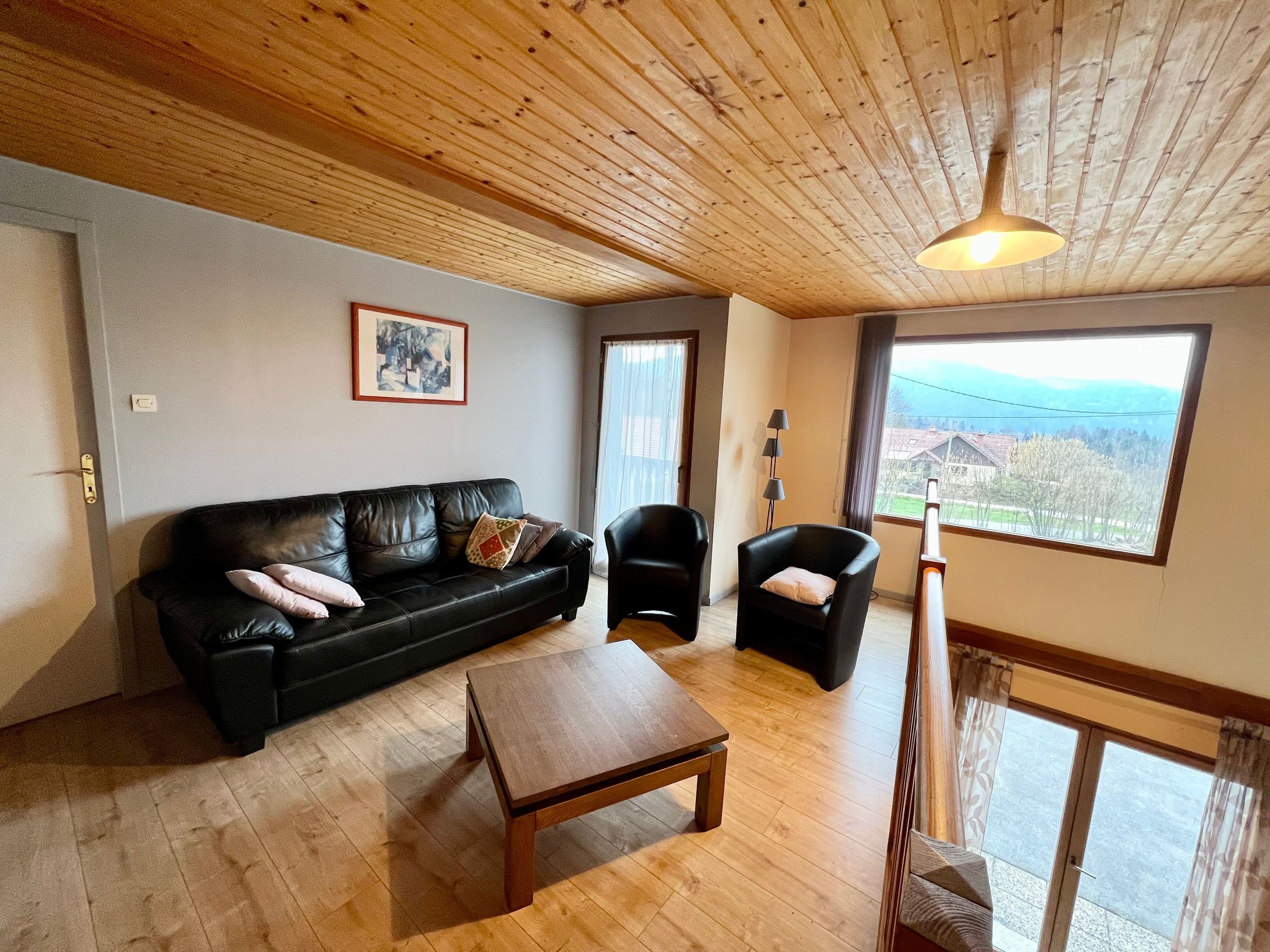 A living room with a wooden ceiling, a black leather sofa, a wooden coffee table, and a view of a landscape through a large window.