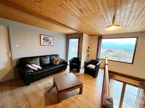 A living room with a wooden ceiling, a black leather sofa, a wooden coffee table, and a view of a landscape through a large window. - Spacious chalet near Gérardmer (Gerbamont)