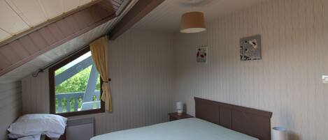 A bedroom with a sloped ceiling, a bed with a light-colored bedspread, a nightstand with a lamp, a window with curtains, and a small table with papers.