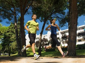 Twee personen joggen op een pad, omringd door bomen en een gebouw op de achtergrond.
