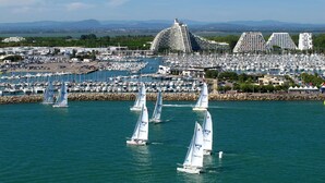 Un puerto deportivo con varios veleros, un edificio grande de diseño singular, y una vista despejada del paisaje circundante.