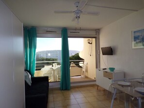 A room with a sofa, a dining table, a TV, and a balcony with a view of buildings. - Studio cabin with pool, close to beach and shops (La Grande-Motte)