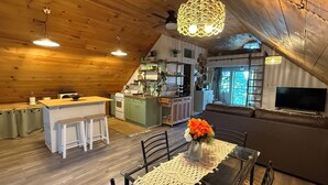 A cozy loft-style living space with a kitchen, dining area, and a large window overlooking greenery.