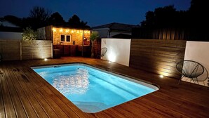 Una piscina rectangular con un muelle de madera, rodeada por una cerca de madera y un pequeño edificio con luces.