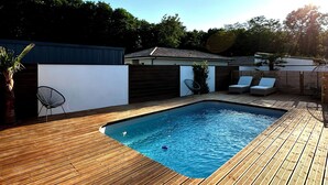 Una terraza de madera con piscina, sillas de descanso y cielo azul despejado.