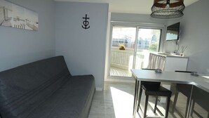 A modern living room with a grey sofa, a dining table with chairs, a pendant light, and a balcony with a view. - Charming Studio Steps from the Beach with Parking and Wi-Fi (La Grande-Motte)