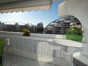 Un balcón con vistas a un moderno complejo de edificios, una planta verde y un toldo color beige.