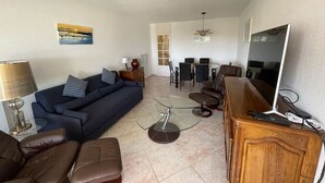 Un salon comprenant un canapé, des fauteuils, une table basse en verre, un téléviseur et un coin repas avec des chaises et une table.
