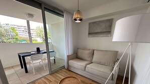 A modern living room with a sofa, a small wooden table, a glass door leading to an outdoor area, and a pendant light. - Luxury A/C Apartment Near Beach and Shops - 2 Rooms, Parking, High-Quality Amenities (La Grande-Motte)