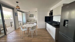 A modern kitchen with a stainless steel refrigerator, white cabinetry, and a dining area with a round table and chairs. - Luxury A/C Apartment Near Beach and Shops - 2 Rooms, Parking, High-Quality Amenities (La Grande-Motte)